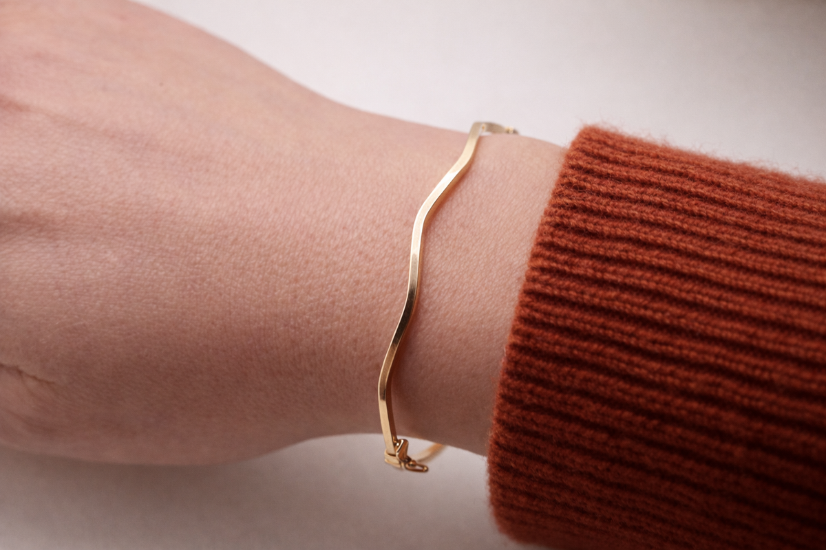 Golvende bangle hoekige vorm 14k goud, gedragen om de pols bij een roestbruine mouw